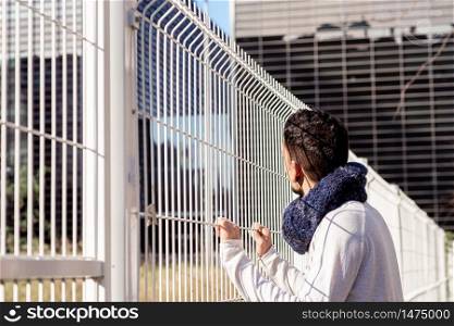 Young male leaning on a metallic fence while looking away to the business buildings