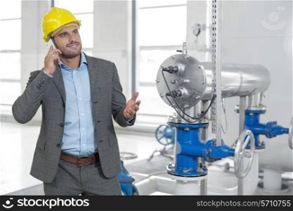 Young male manager in hard hat using cell phone by machinery in industry
