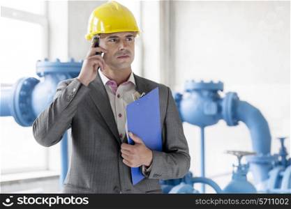 Young male manager with clipboard using cell phone in industry