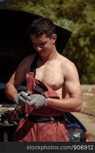 Young Male Mechanic in red overalls and a naked torso stands at a car with an open hood. Young succesful professional car repairman in flannel and overalls standing by automobile under repair