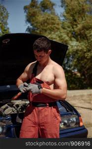 Young Male Mechanic in red overalls and a naked torso stands at a car with an open hood. Young succesful professional car repairman in flannel and overalls standing by automobile under repair