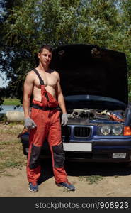 Young Male Mechanic in red overalls and a naked torso stands at a car with an open hood. Young succesful professional car repairman in flannel and overalls standing by automobile under repair