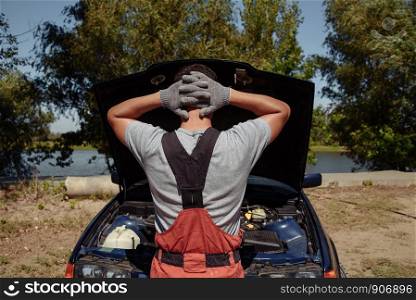 Young Male Mechanic is standing at a car with an open hood and does not know what to do. Mechanic standing in front of the open hood and repairing the car