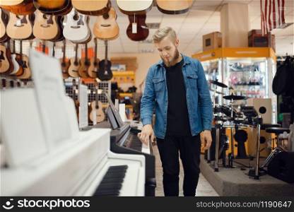 Young male musician poses at the piano in music store. Assortment in musical instruments shop, keyboardist buying equipment, pianist in market. Young musician poses at the piano in music store