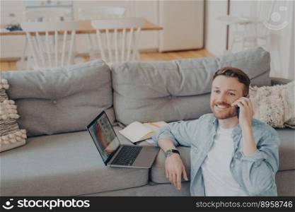 Young male office worker working from home, talking on phone with his co-worker about projects status, preparing presentation with gathered data, resting hand on couch while sitting on floor. Young male office worker working from home talking on phone with his co-worker