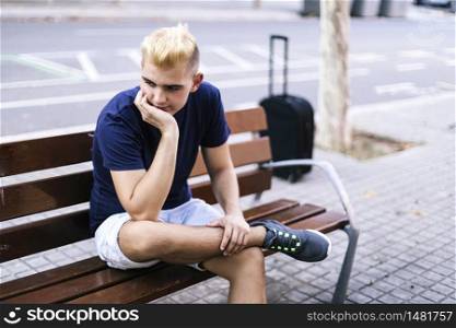 Young male sitting on a bench in the street with his hands on his face worried and sad