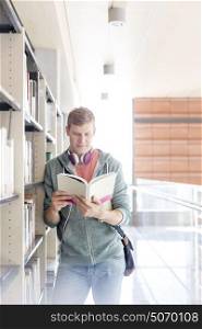 Young male student reading book by shelf at university library