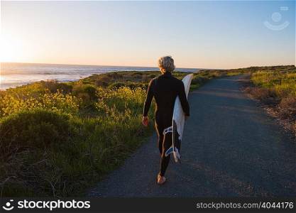 Young male surfer at sunrise carrying surfboard on rural road at coast