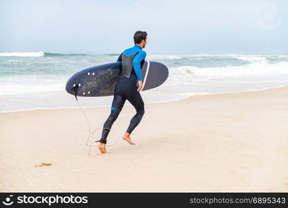 Young male surfer wearing wetsuit, holding surfboard under his arm, running on beach after morning surfing session.