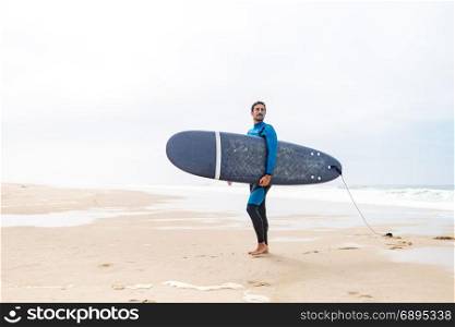 Young male surfer wearing wetsuit, holding surfboard under his arm, standing on beach after morning surfing session.