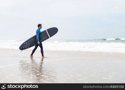 Young male surfer wearing wetsuit, holding surfboard under his arm, walking on beach after morning surfing session.