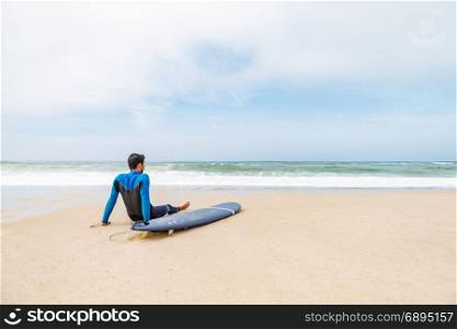 Young male surfer wearing wetsuit, sitting beside his surfboard on beach after morning surfing session.