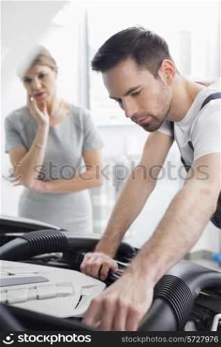 Young male technician examining car engine with worried customer in background at workshop