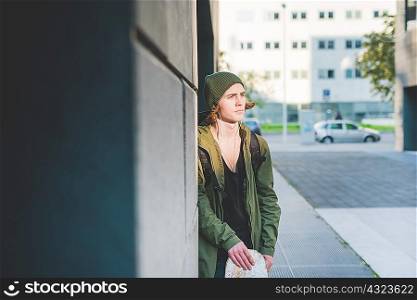 Young male urban skateboarder leaning against wall listening to earphone music