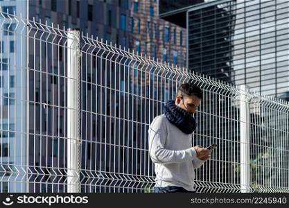Young male wearing scarf leaning on metallic fence while using smartphone outdoors
