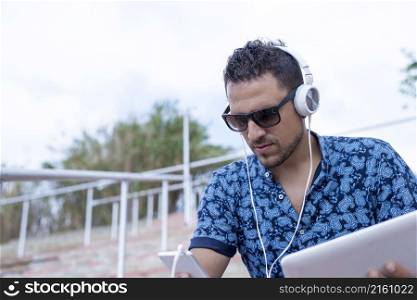 Young male with headphones and sunglasses sitting on stairs outdoors using tablet