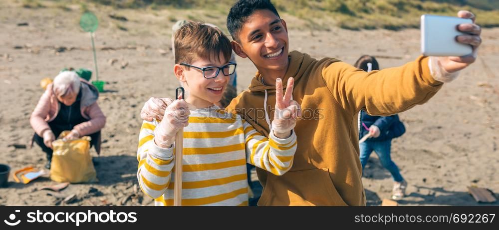 Young man and boy taking selfie with mobile to group of volunteers cleaning the beach. Selective focus on guy and boy in foreground. Young man and boy taking selfie to group of volunteers cleaning beach