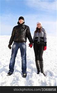 young man and girl standing on snowy area, holding for hands and smiling, looking at camera