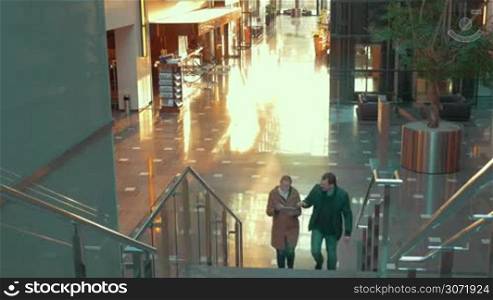 Young man and woman going upstairs in shopping centre. They talking and using tablet PC. Modern interior, sunlight reflecting on tiled floor