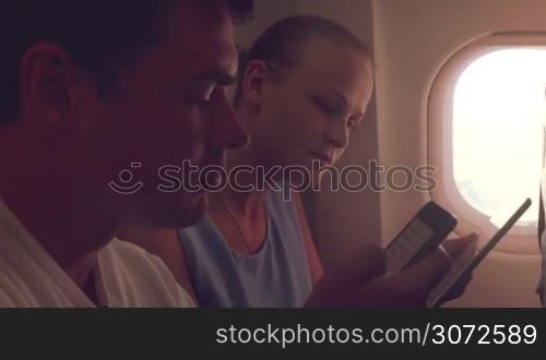 Young man and woman having a conversation in plane while she using tablet computer and he typing text message on smart phone