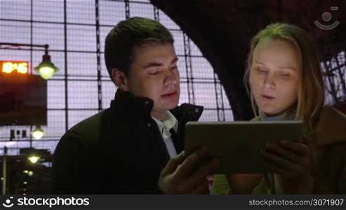 Young man and woman having vivid conversation while using tablet computer at the railway station in the evening