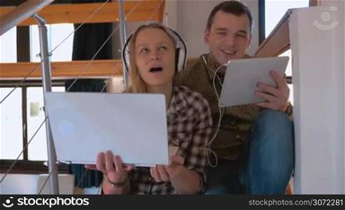 Young man and woman in headphones listening to music and dancing at home. They using laptop and touch pad sittig on the stairs