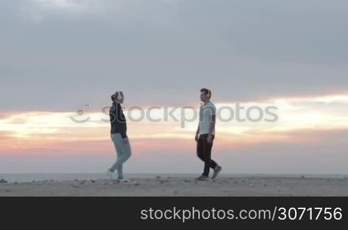 Young man and woman in wireless headphones doing physical exercises on the beach at sunset