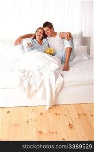 Young man and woman lying down in white bed watching television and eating crisps