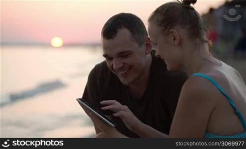 Young man and woman on the beach at sunset looking through the photos on touch pad, talking and laughing