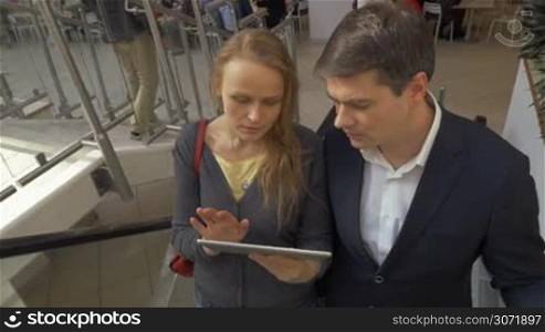 Young man and woman riding up on escalator in shopping center and discussing some business matters using tablet computer