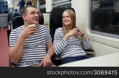 Young man and woman talking and drinking tea or coffee while traveling by train in the evening