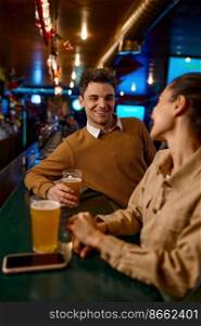 Young man and woman talking while enjoying drinks together at sport pub. Friendship, flirting and relationship. Focus on smiling guy at bar counter. Man and woman talking while enjoying drinks together