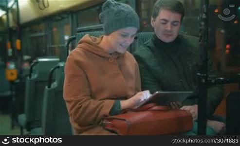 Young man and woman traveling by bus in the evening city. They using touch pad and talking during the ride