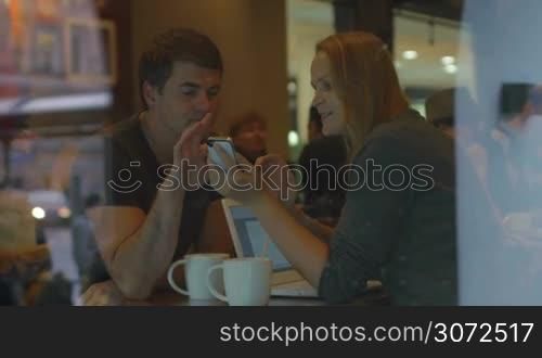 Young man and woman watching something using cell phone and discussing it during tea break in cafe. Street with people reflecting in glass