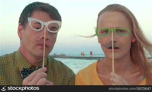 Young man and woman with hipster style paper glasses and moustache having fun while making goofy faces outdoor by the sea. Trendy style