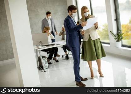 Young man and woman with protective facial masks discussing with paper in hands indoors in the office with young people works behind them