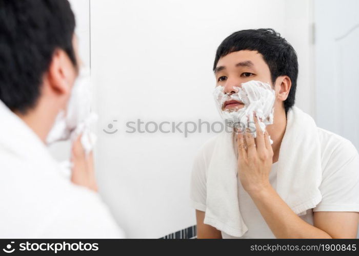 young man applying foam cream on face before shaving in the bathroom mirror