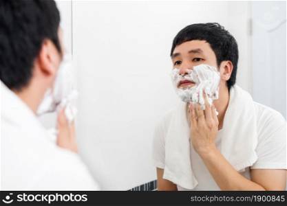 young man applying foam cream on face before shaving in the bathroom mirror