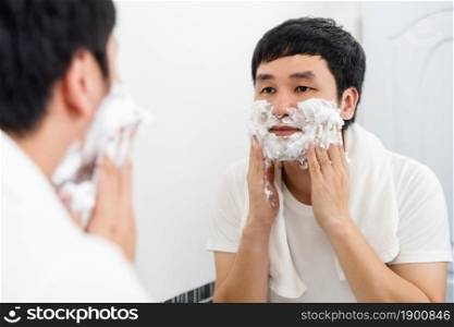 young man applying foam cream on face before shaving in the bathroom mirror