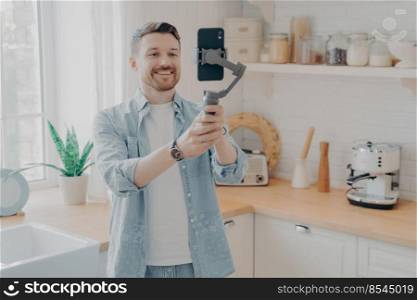 Young man blogger recording video blog or live streaming with smartphone and gimbal indoors standing in kitchen background. Social media vlogger shooting vlog, streaming online podcast on mobile phone. Young man blogger recording video blog with mobile phone at home