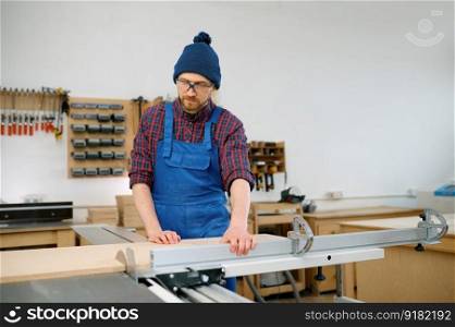 Young man carpenter working on circular saw and engaged in making wooden blank at workshop. Man working on circular saw and engaged in making wooden blank at workshop