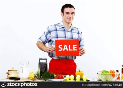 Young man cooking fresh meal at home and holding sale sign