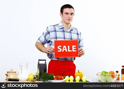 Young man cooking fresh meal at home and holding sale sign