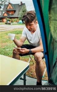 Young man drinking a coffee sitting at front of tent in the morning. Teenager enjoying free time during weekend trip in summer. Young man drinking a coffee sitting at front of tent in the morning