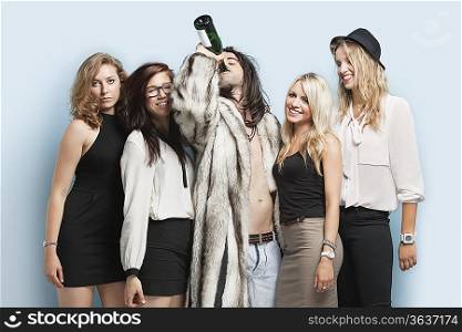 Young man drinking beer while standing with female friends against light blue background