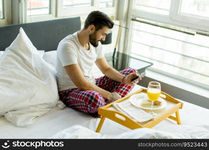 Young man eating breakfast in bed whilst using mobile phone