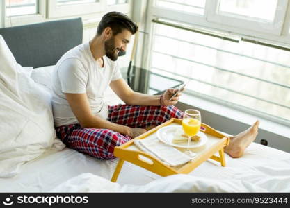 Young man eating breakfast in bed whilst using mobile phone