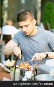 Young man eating double cheeseburger with fork and knife in outdoor restaurant