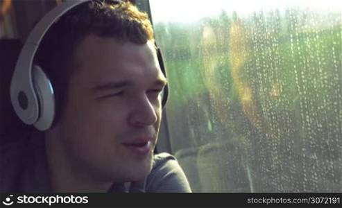Young man enjoying his train ride. He listening to music in headphones, singing and dancing while looking out the window