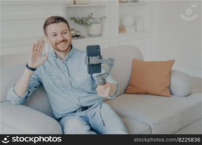 Young man enjoying video chat with friends , looking at phone screen and waving with hand, cheerful bearded man holding smartphone with gimbal stabilizer handheld while sitting on sofa in living room. Young man enjoying video chat with family on cellphone while sitting on sofa in living room